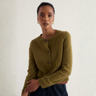 A woman wearing a cropped olive green knit cardigan with dark buttons, paired with dark navy trousers, standing against a plain off-white wall.
