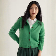 Load image into Gallery viewer, A woman wearing a bright green knit cardigan with brown buttons over a white collared shirt and a dark green skirt, standing against a plain off-white background.
