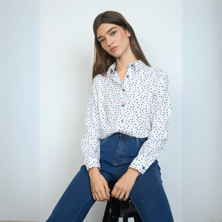Woman wearing a white blouse with a floral pattern and blue jeans sitting on a stool against a light gray background