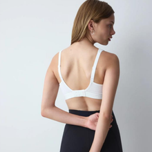 Load image into Gallery viewer, A woman shown from the back wearing a white open-back bralette with textured mesh panels and thin straps, paired with black bottoms, against a plain light gray studio background.
