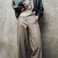 Load image into Gallery viewer, A person wearing wide-leg taupe trousers, a gray graphic t-shirt, and a dark green cardigan, standing against a textured concrete wall.
