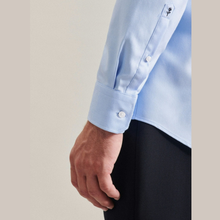 Load image into Gallery viewer, Close-up of a light blue long-sleeve dress shirt worn by a model, showing the buttoned cuff and part of the pocket, paired with dark trousers against a neutral beige background.
