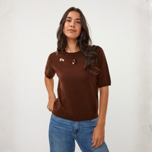 Load image into Gallery viewer, A woman modeling a short-sleeve brown knit top with small metallic decorative embellishments on the chest, paired with blue jeans, against a plain white studio background.
