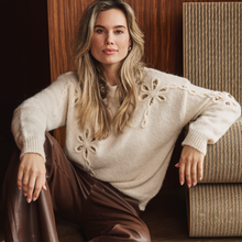 Load image into Gallery viewer, A woman wearing a cream-colored knit sweater with textured floral details and brown leather pants, sitting in a warm, rustic interior next to a woven chair.
