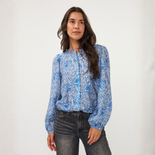 Load image into Gallery viewer, A woman modeling a blue and white patterned long-sleeve blouse with puffed shoulders, paired with dark gray jeans, against a plain white studio background.
