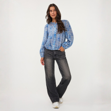 Load image into Gallery viewer, A woman modeling a blue and white patterned long-sleeve blouse with dark gray jeans and white sneakers, standing against a plain white studio background.
