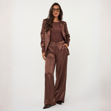 Load image into Gallery viewer, A model with brown hair stands against a light gray studio background wearing a coffee coloured long-sleeve blouse paired with brown wide-leg trousers.

