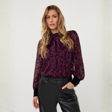 Load image into Gallery viewer, A woman modeling a long-sleeve sheer lace blouse in deep purple with a tonal floral pattern, paired with black pants, against a plain off-white studio background.
