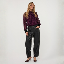 Load image into Gallery viewer, A woman modeling a long-sleeve purple and black patterned blouse with black faux-leather wide-leg pants and dark pointed-toe heels, standing against a plain light gray studio background.
