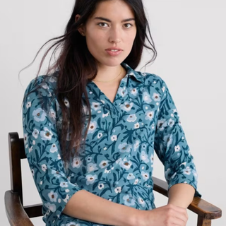 A woman wearing a blue floral button-up shirt, seated in a wooden chair against a light gray background.