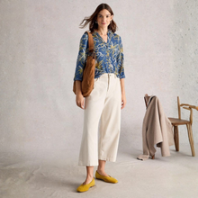 Load image into Gallery viewer, Woman wearing a blue floral blouse, white pants, and yellow shoes, standing in a minimalistic room.
