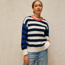 Load image into Gallery viewer, A woman wearing a color-blocked knit sweater with navy, white, and royal blue horizontal stripes, featuring a red ribbed crew neck and white ribbed cuffs and hem, paired with light blue jeans against a textured beige wall.
