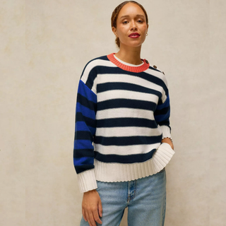 A woman wearing a color-blocked knit sweater with navy, white, and royal blue horizontal stripes, featuring a red ribbed crew neck and white ribbed cuffs and hem, paired with light blue jeans against a textured beige wall.