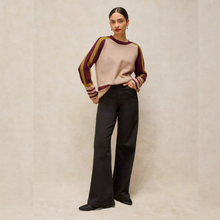Load image into Gallery viewer, A woman stands against a textured beige studio backdrop wearing a knit sweater with a beige body and dark burgundy sleeves, paired with black wide-leg trousers and black pointed shoes.

