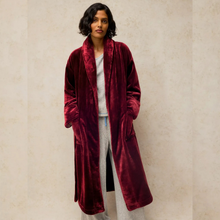 Load image into Gallery viewer, A woman wearing a plush, long burgundy velvet robe over a light gray lounge set, standing against a textured beige wall.
