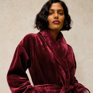 A woman wearing a plush, deep burgundy velvet bathrobe against a textured beige background.