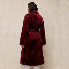 Load image into Gallery viewer, A woman viewed from the back wearing a long burgundy velvet robe with a matching waist tie, standing against a textured beige wall.
