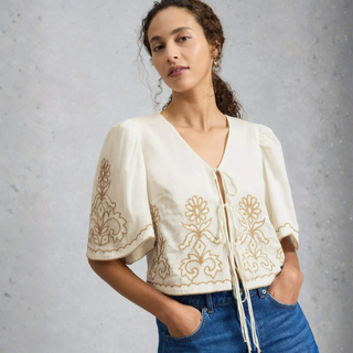 Woman wearing a cream embroidered top with blue jeans against a gray background