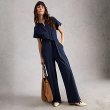 Load image into Gallery viewer, Woman wearing a navy blue jumpsuit holding a brown bag against a gray background
