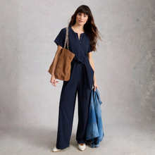 Load image into Gallery viewer, Woman in navy jumpsuit holding a brown bag and denim jacket against a gray background
