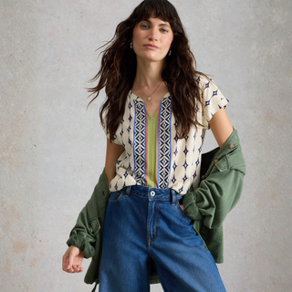 Woman wearing a patterned top, green jacket, and blue jeans against a neutral background