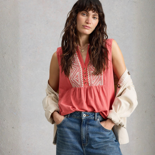 Woman wearing a pink embroidered blouse with a beige cardigan and blue jeans against a gray background