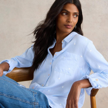 Load image into Gallery viewer, Woman wearing a light blue shirt sitting on a wooden chair against a beige background
