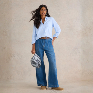 Woman wearing a light blue shirt and blue jeans, holding a patterned handbag against a beige background