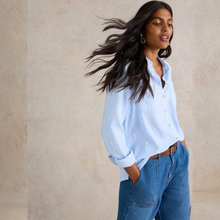 Load image into Gallery viewer, Woman wearing a light blue shirt and blue jeans against a beige wall
