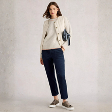 Load image into Gallery viewer, A woman stands against a light textured wall wearing a cream cable-knit cardigan over a cream sweater, dark navy cropped pants, and brown lace-up sneakers. She is holding a small dark blue bag.
