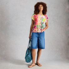 Load image into Gallery viewer, A woman stands against a textured gray studio backdrop wearing a bright multicolor floral and patterned short-sleeve top, blue denim shorts, and tan sandals, while holding a blue patterned tote bag.
