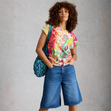Load image into Gallery viewer, A woman with curly brown hair stands against a textured light gray wall, wearing a bright multicolor floral short-sleeve top, blue denim culottes, and carrying a teal patterned shoulder bag.
