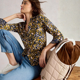 Woman sitting on a chair wearing a patterned blouse and jeans, with a bag beside her.