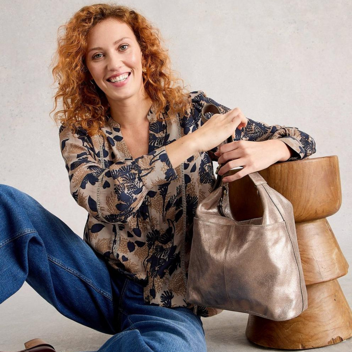 Woman holding a metallic handbag sitting on a wooden stool against a plain background