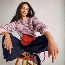 Load image into Gallery viewer, Woman sitting on a stool wearing a colorful sweater, jeans, and patterned shoes with a red bag.
