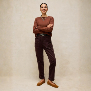 A woman stands against a warm beige textured backdrop wearing a rust-brown long-sleeve knit top, dark brown corduroy trousers, a black belt, and brown flats.