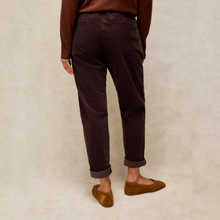 Load image into Gallery viewer, A person shown from the back wearing dark plum corduroy trousers, a brown sweater, and brown suede flats, standing against a textured beige background.
