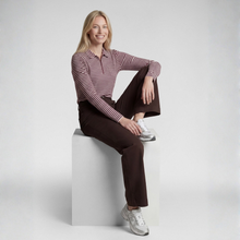 Load image into Gallery viewer, A woman sits on a white cube in a studio setting, wearing a long-sleeve polo shirt with thin red and white horizontal stripes, dark brown pants, and white sneakers.
