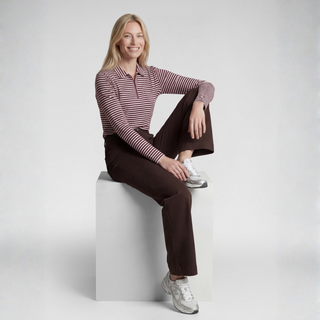 A woman sits on a white cube in a studio setting, wearing a long-sleeve polo shirt with thin red and white horizontal stripes, dark brown pants, and white sneakers.
