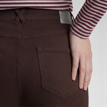 Load image into Gallery viewer, Close-up of a person wearing dark brown jeans, showing the back pocket, waistband, and hand resting on the thigh, with a striped long-sleeve top partially visible.
