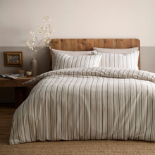 Load image into Gallery viewer, A bed dressed in a striped duvet cover and matching pillow shams in beige, gray, and muted blue-gray tones, styled in a bedroom with a wooden headboard, bedside table, and neutral wall.
