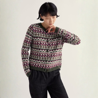 A model wearing a long-sleeve knit cardigan sweater with a geometric Fair Isle pattern in green, pink, and beige, paired with dark charcoal pants, against a plain off-white background.