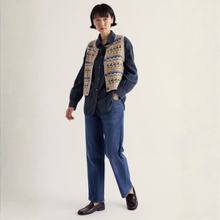 Load image into Gallery viewer, A model stands against a plain off-white studio background wearing a dark blue button-up shirt, blue jeans, dark brown loafers, and a beige knit vest with a colorful geometric pattern.
