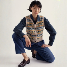 Load image into Gallery viewer, A model crouches in a studio setting wearing a patterned Fair Isle knit cardigan vest over a dark blue button-up shirt, paired with blue jeans and dark brown leather loafers.
