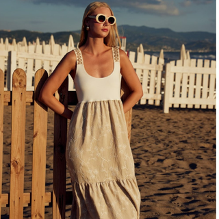 Woman in a white top and beige dress standing on a beach with a wooden fence and white picket fence in the background.